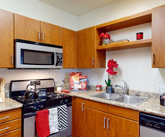 A cozy kitchen with red decorations, red flowers, a cookbook, stainless steel appliances, marble countertops with lots of room, and large wooden cabinets at Vesada Apartment Homes. 