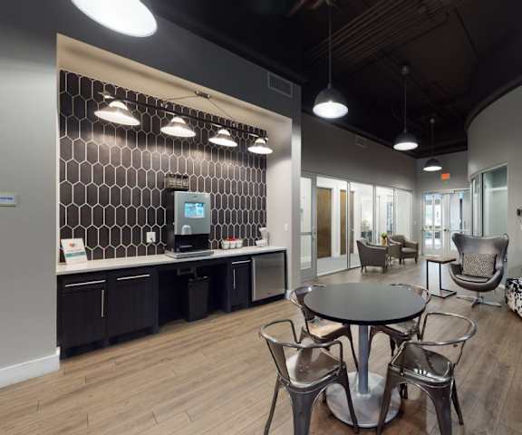 The community lounge with chairs, tables, plush chairs, a coffee machine, toaster, fridge, marble counters, and barstool chairs at Lofts at Watters Creek Apartments.