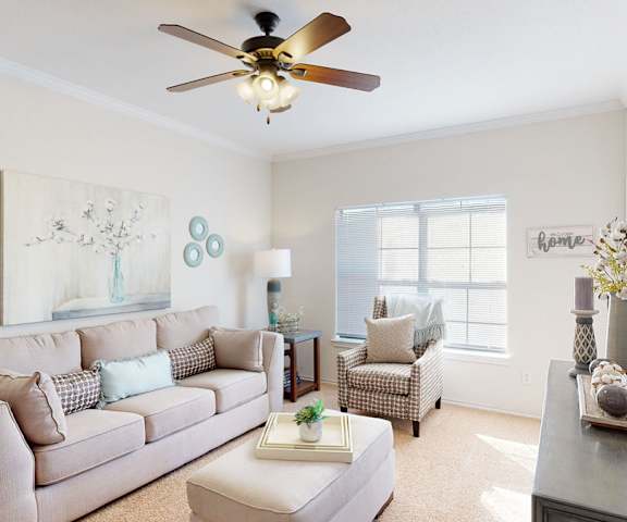 The spacious living room with a tan couch, throw pillows, large windows, a ceiling fan, wall art, plush chair, coffee table, decorations, large windows and a mirror at Stoneleigh on May Apartment Homes. 