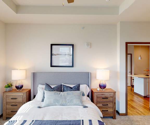 A spacious bedroom with a queen-size bed with a white and gray bedspread, a dresser, wall art, decorations, lamps, a closet, and nightstands at Van Buren East Apartment Homes. 