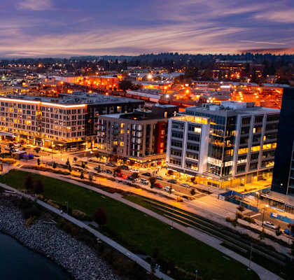 A cityscape at dusk with buildings and lights along a river.