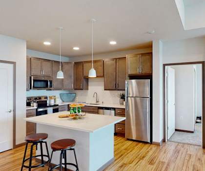 A large, modern kitchen with stainless steel appliances, white marble countertops with lots of room, a kitchen island with barstools, and large medium wooden cabinets at Van Buren West Apartment Homes. 