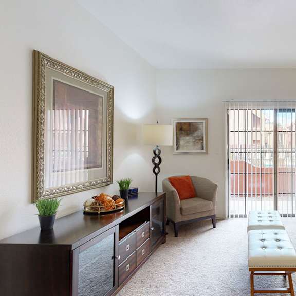 A living room with a couch, some artwork on the wall, a plush chair, some footstools, and a door to a balcony at Atrium Court.