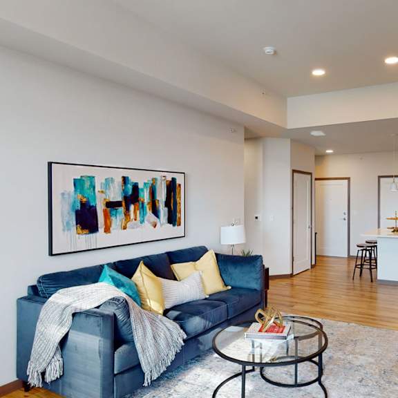 The spacious living room with a blue, velvet couch, plush chairs, coffee table, overhead lighting with a ceiling fan, decorations, a TV, and view of the kitchen at Van Buren East Apartment Homes. 