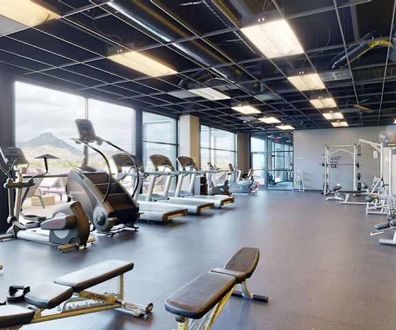 A spacious gym with cardio machines, large windows, a yellow accent wall, TVs, and weights at the Trend at 51 Apartment Homes. 