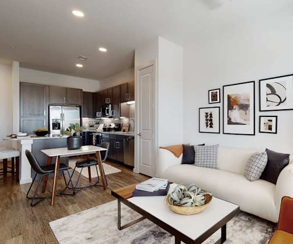The large living room with a white couch, plush chairs, coffee table, decorations, a TV, and view of the dining room and the kitchen with marble counters and black appliances at Magnolia on the Green Apartments. 