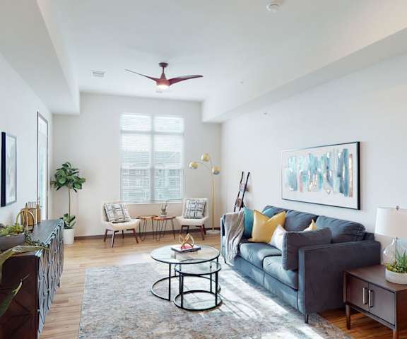 The spacious living room with a dark gray couch, dark wood drawers, ceiling fan, houseplants, wall artwork, yellow throw pillows, large windows, a ceiling fan, wall art, plush chairs, coffee table, decorations, a TV, and windows with blinds at Van Buren West Apartment Homes. 