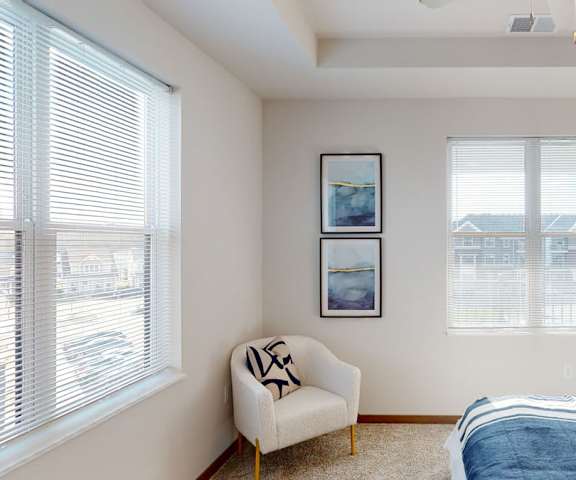 A spacious bedroom with a queen-size bed with a blue bedspread, a dresser, wall art, lamps, white plush chair, decorations, two windows, and nightstands at Van Buren West Apartment Homes. 