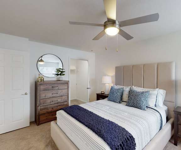 A spacious bedroom with a queen-size bed with a white and blue bedspread, a mirror, a ceiling fan, a dresser, decorations, closet, and nightstands at Villas at Preston Lakes Apartment Homes. 