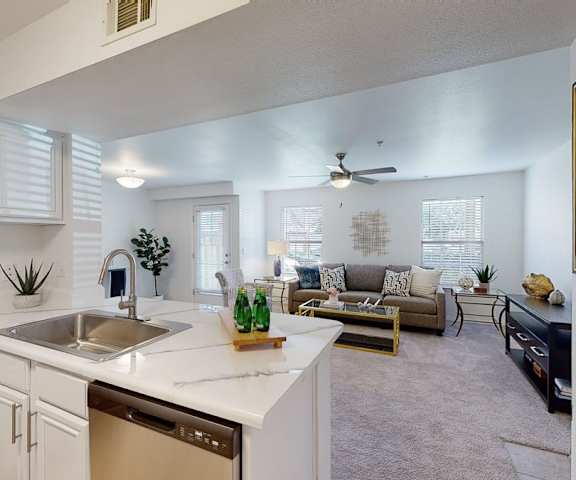 The spacious living room with a couch, plush chairs, coffee table, overhead lighting with a ceiling fan, decorations, a TV, and view of the dining room with a table, chairs, and place settings for four, and the kitchen with white counters and stainless-steel appliances at Villas at Preston Lakes Apartment Homes. 