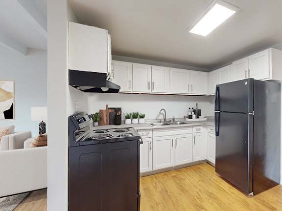A black refrigerator stands in a kitchen with white cabinets and a wooden floor.