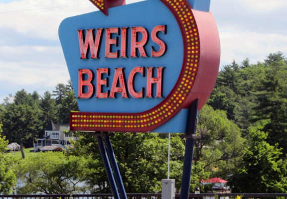 A sign that says Weirs Beach stands in front of a green landscape.