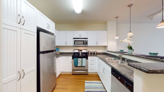 A large, modern kitchen with stainless steel appliances, dark granite countertops with lots of room, a white orchid, two place settings at the bar, modern lighting, and large white wooden cabinets at Vesada Apartment Homes. 