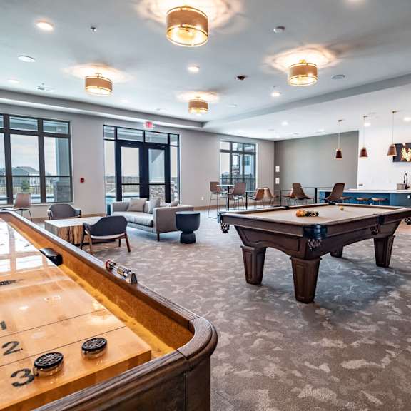 A game room with a pool table, arcade games, chairs, tables, kitchenette with a TV, and a shuffleboard at Van Buren West Apartment Homes.