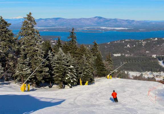 A skier is seen on a snowy slope with a lake and mountains in the background.