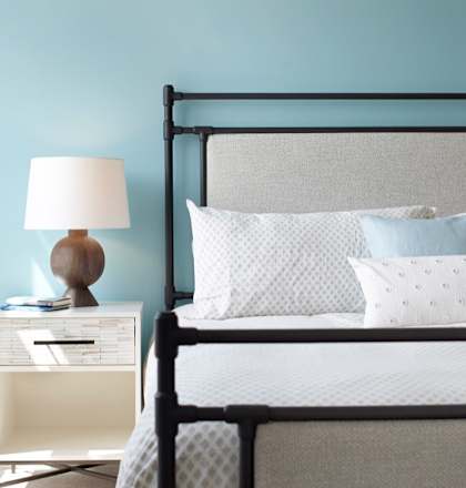 A bed with a white comforter and two pillows sits next to a nightstand with a lamp on it.
