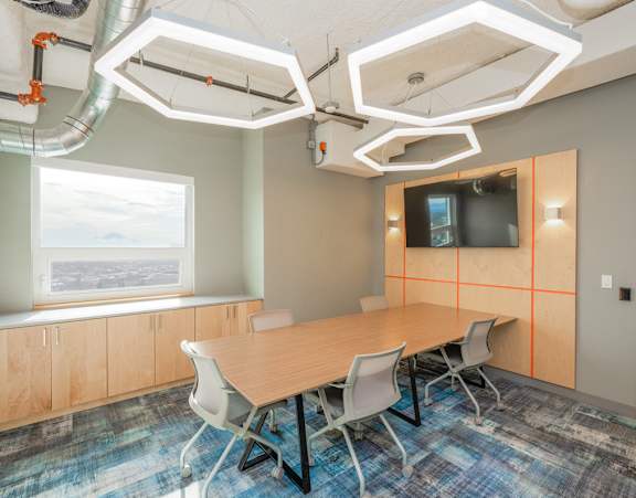 A conference room with a long table and chairs, a television on the wall, and a window with a view of the sky.