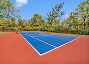 Thumbnail 5 of 5 - A tennis court with a blue border and red surface surrounded by trees.