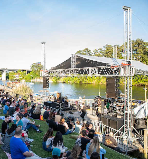 A large crowd of people are sitting on the grass watching a concert by a lake.