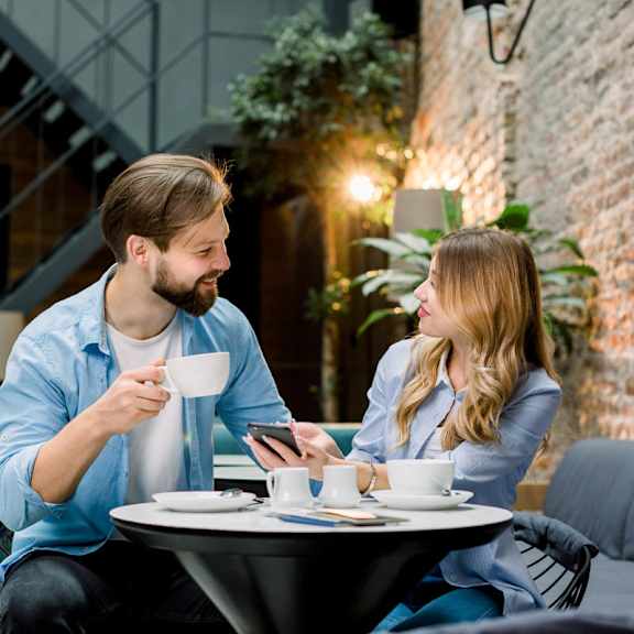  A man and a woman are sitting at a table in a cafe, with the man holding a cup and the woman looking at her phone.