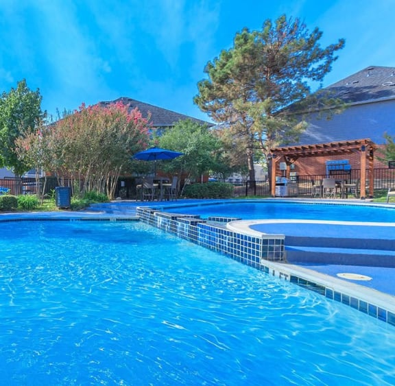 A large blue swimming pool surrounded by trees and a building in the background. at Villas of Sorrento Apartments, Dallas