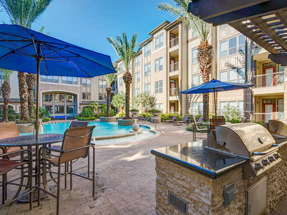 A large swimming pool surrounded by a stone patio and umbrella tables.