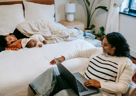 A woman is sitting on a bed with a laptop, while a child sleeps next to her. at Pine Creek Apartments, Houston Texas