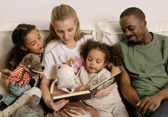 A family of four is sitting on a couch, reading a book together.