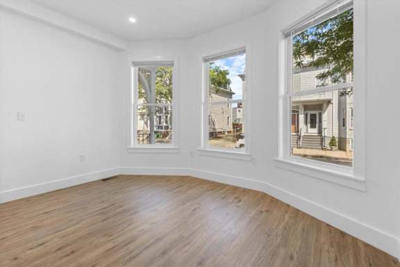 A room with white walls and wooden flooring, featuring two windows with views of the outside.
