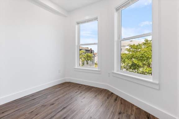 A room with white walls and wooden flooring, featuring two windows with a view of greenery and buildings outside.