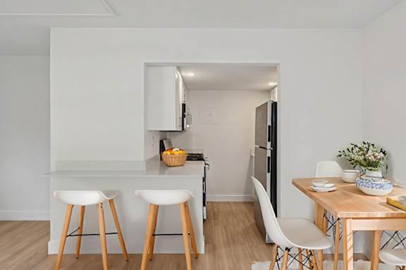 A kitchen with white walls and wooden floors, featuring a bar area with two stools and a countertop with a bowl of fruit on it.