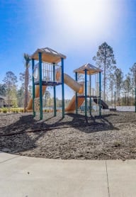 Bright playground at Treeline Trails with a yellow slide, swings, and benches, surrounded by tall pines under a sunny blue sky.