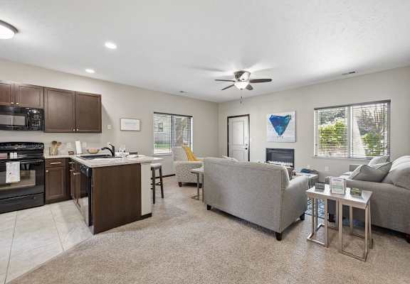 Staged open living room and kitchen with carpet, ceiling fan, fireplace, gray seating, and breakfast bar.