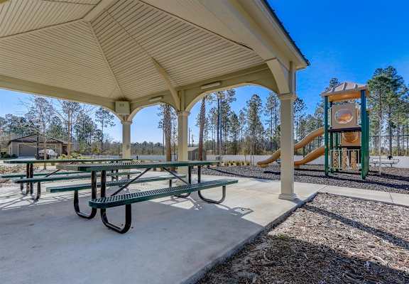 Covered picnic area at Treeline Trails with green tables overlooking a playground with a yellow slide under a bright blue sky.