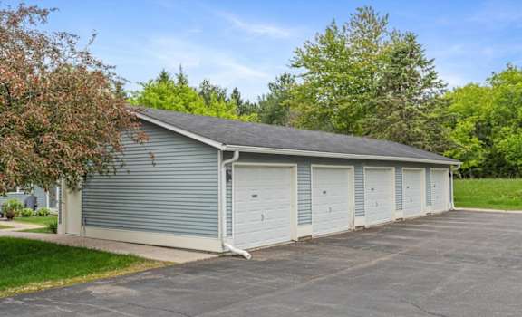 A two-car garage with a grey roof and white doors is surrounded by a paved driveway and greenery.