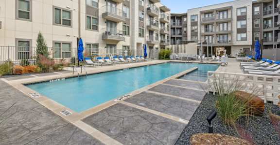 A large swimming pool surrounded by a stone patio and lounge chairs in front of apartment buildings.