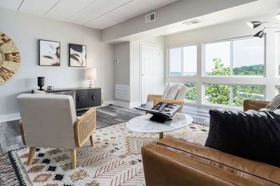 A living room with a brown leather couch, a wooden chair, a round rug, and a large window with a view of trees.  at The Heights Apartments, Cincinnati, 45220