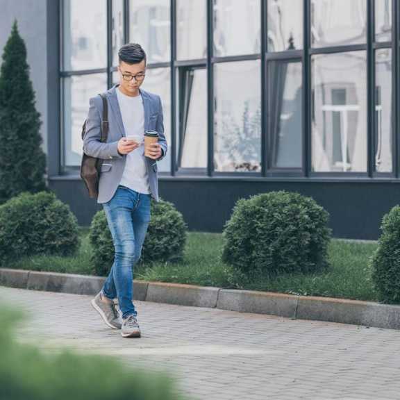 A man in a grey blazer and blue jeans is walking while looking at his phone.