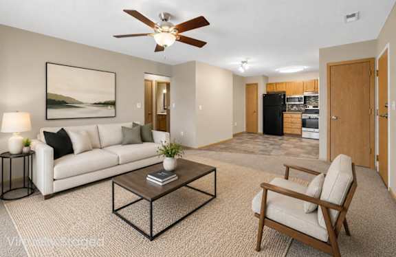 Virtually staged living area with a sofa, coffee table, accent chair, ceiling fan, and an open connection to the kitchen.