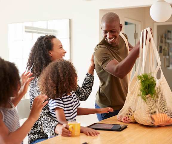 A man is holding a bag with vegetables in it while two children look on.
