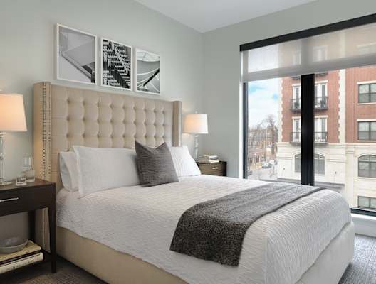 A bedroom with a large bed and a window overlooking a cityscape. at The Elle on Lockwood Apartments, Webster Groves, MO, 63119
