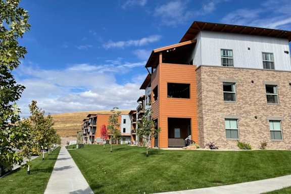 A modern building with a mix of stone and wood paneling is surrounded by a well-kept lawn and a clear blue sky.