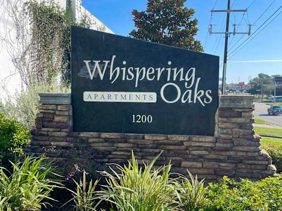 A sign for Whispering Oaks Apartments stands in front of a tree.
