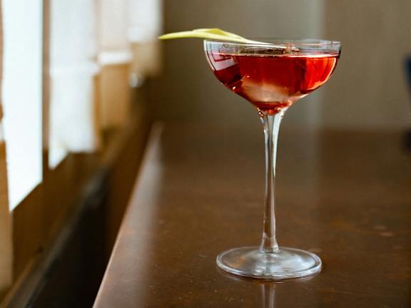 A red cocktail in a wine glass with a lemon twist on the rim.