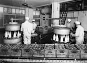 Thumbnail 7 of 8 - Three workers in a factory setting are surrounded by large containers and machinery.