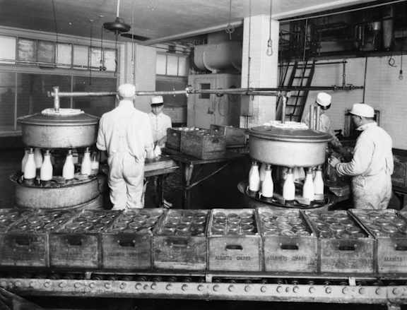 Three workers in a factory setting are surrounded by large containers and machinery.