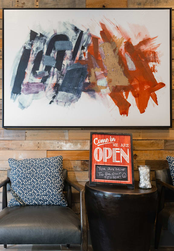 A painting hangs on a wooden wall above a black leather couch with a sign that says "Come in, open.".