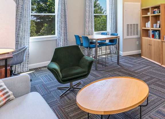 The Residences at Crosspoint cyber lounge in Lowell, MA includes a green accent wall with a mounted flat-screen TV, a wooden storage unit with open shelving, a dark green swivel chair, a round wood coffee table, and a high-top table with blue chairs by two curtained windows overlooking trees.