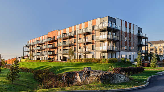 A modern multi-story building with balconies is surrounded by a grassy area and a small waterfall.