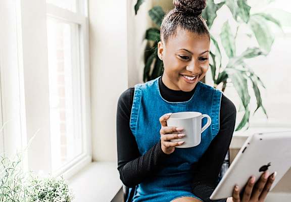 a woman sitting on a couch with a cup of coffee looking at a tablet computer